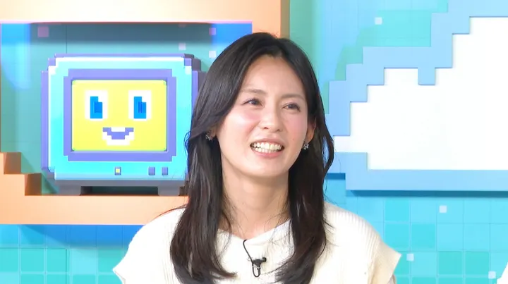 Yuika Motokariya (本仮屋ユイカ) smiling in a white knit top against the colorful Uraaka Uraraka studio backdrop.