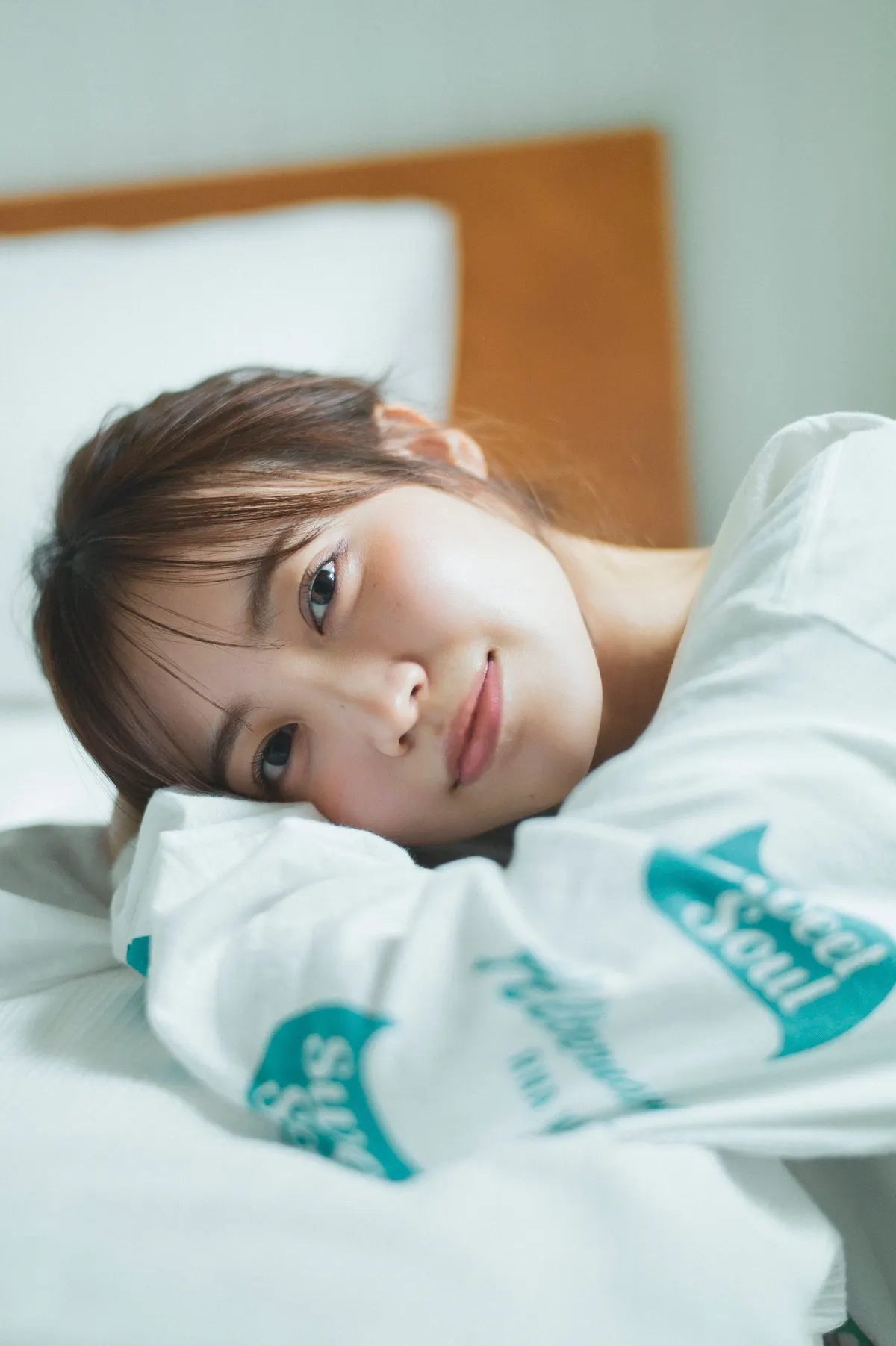 Rina Matsuda resting her head on white bedding in a white printed sweatshirt.