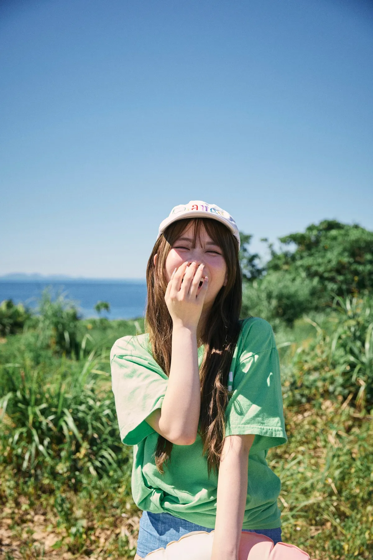 Sakura (さくら) laughing with a hand near her mouth, wearing a green T-shirt and pale cap with vegetation and the sea behind her.