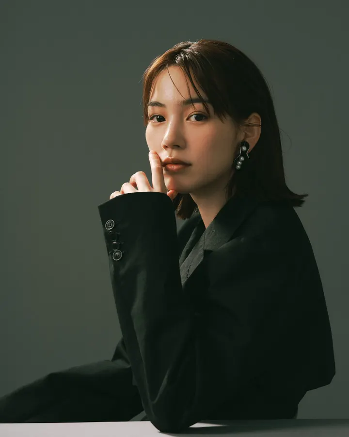Non in a black jacket with one finger beside her chin against a dark gray background.
