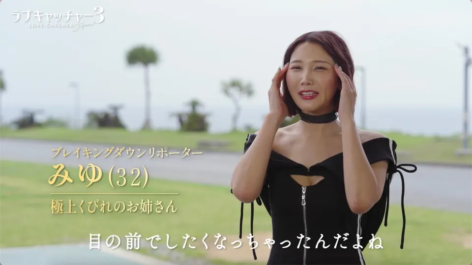 Miyu Kojima (小島みゆ) with short reddish-brown hair, wearing a black off-shoulder outfit and raising her hands beside her face in a Love Catcher Japan 3 interview still.